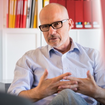 Ein mittelalter Mann mit Brille sitzt vor einem Schrank mit juristischer Literatur